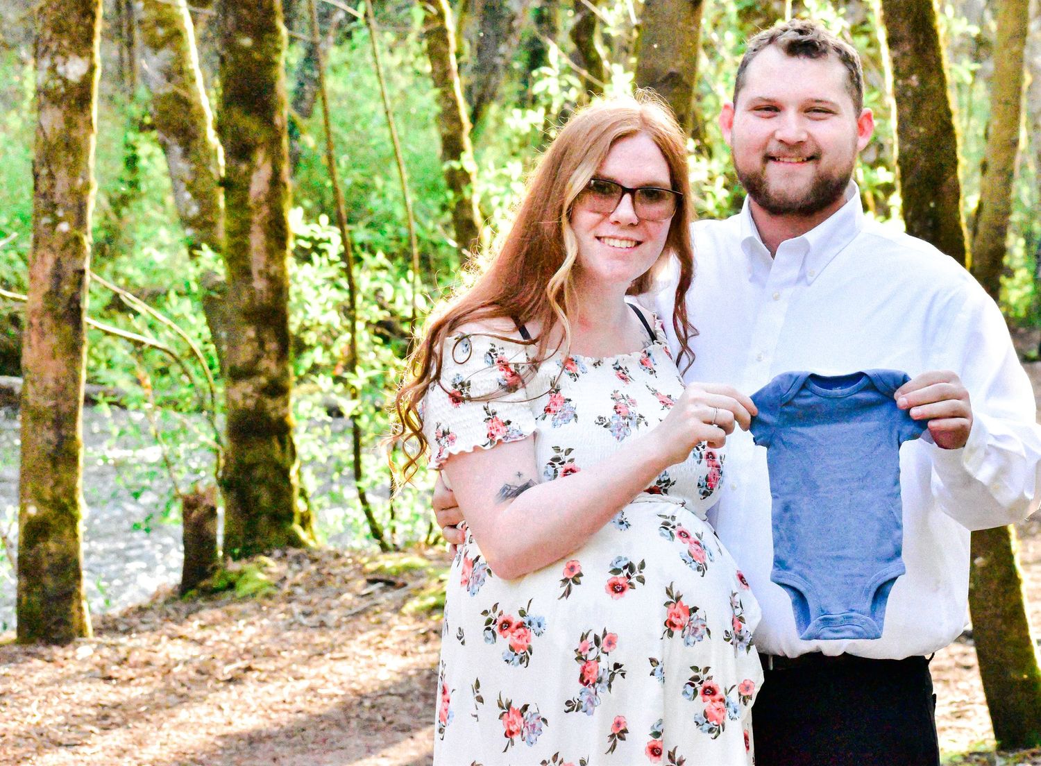 Maternity photo featuring a smiling couple in a peaceful forest setting. The expecting mother wears a floral dress, cradling her bump while