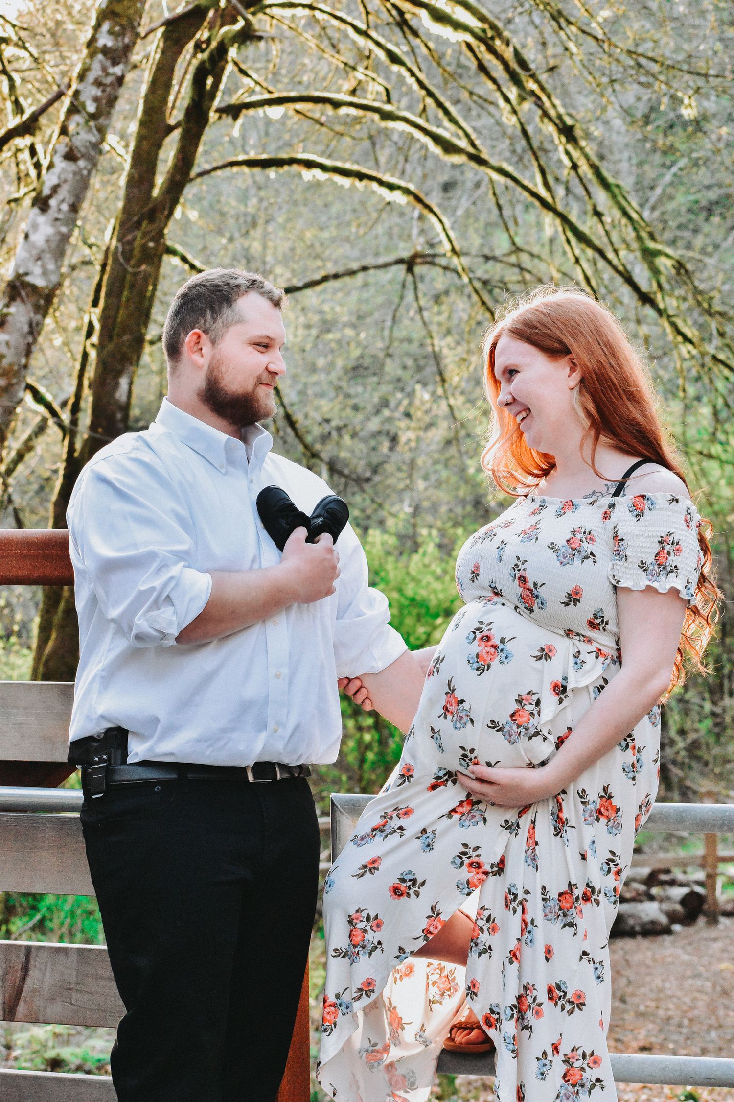 Maternity photo featuring a smiling couple in a peaceful forest setting. The expecting mother wears a floral dress, cradling her bump.