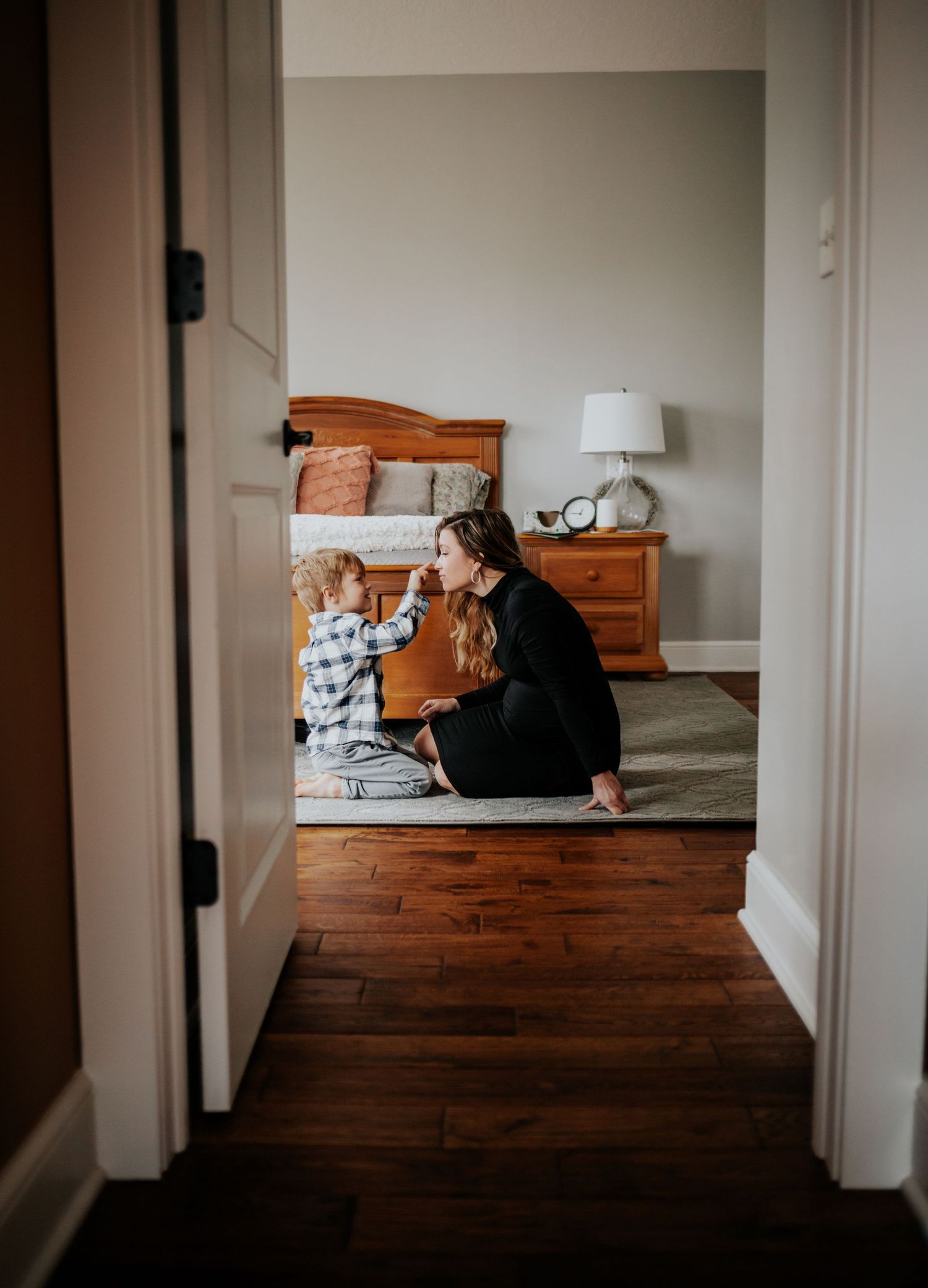 A child and adult sit together on a bedroom floor viewed through a doorway showing hardwood floors and natural lighting.