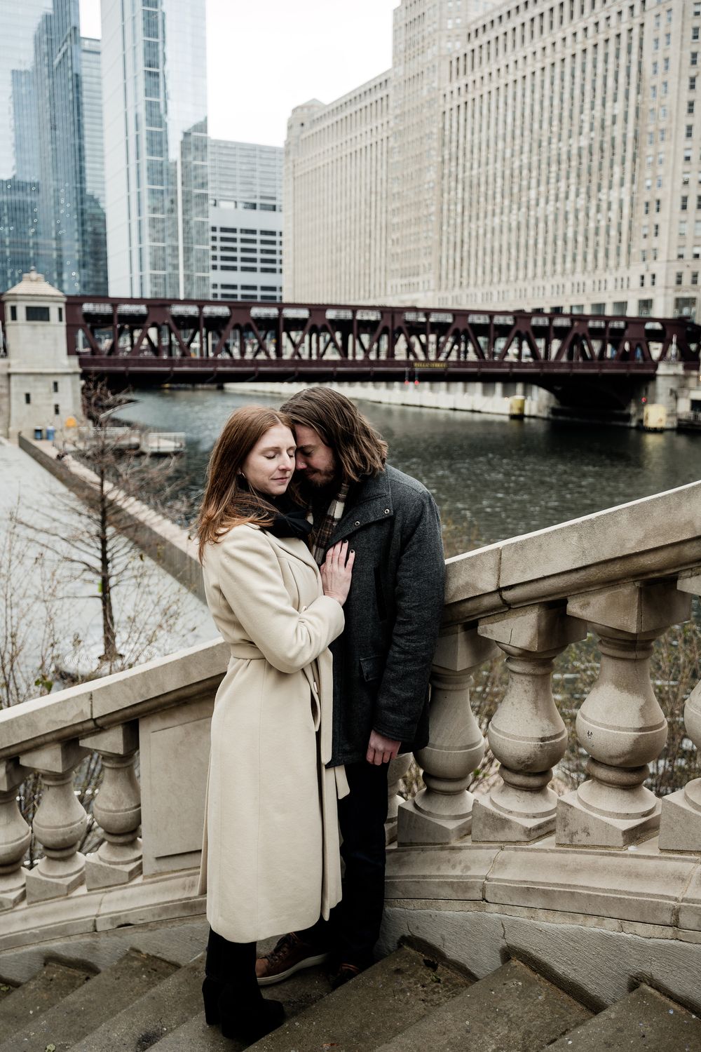A couple embraces on stone steps overlooking a river and Chicago's architectural landmarks.