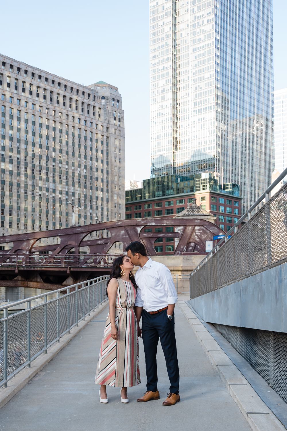 A couple shares an intimate moment on an urban bridge walkway with Chicago's skyline in the background.