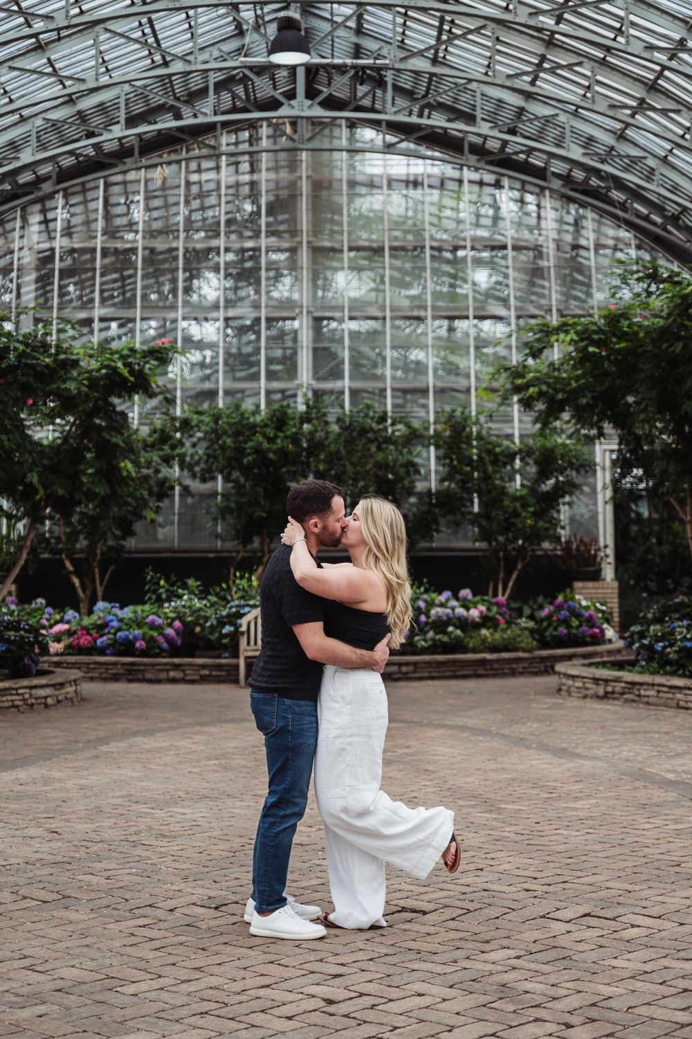A couple embraces in a greenhouse conservatory with glass ceiling and colorful flower gardens.