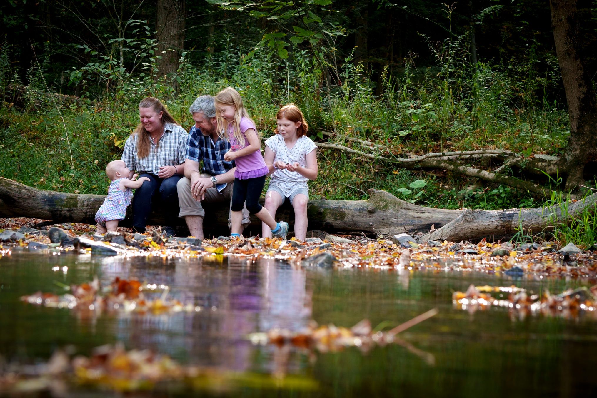 Family group sitting by a pond surrounded by autumn leaves in a forest setting.