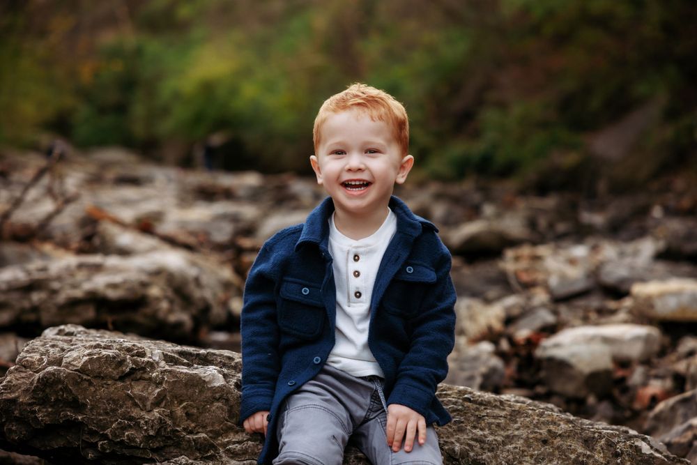 A happy child in a blue jacket sitting on rocky terrain near a stream.