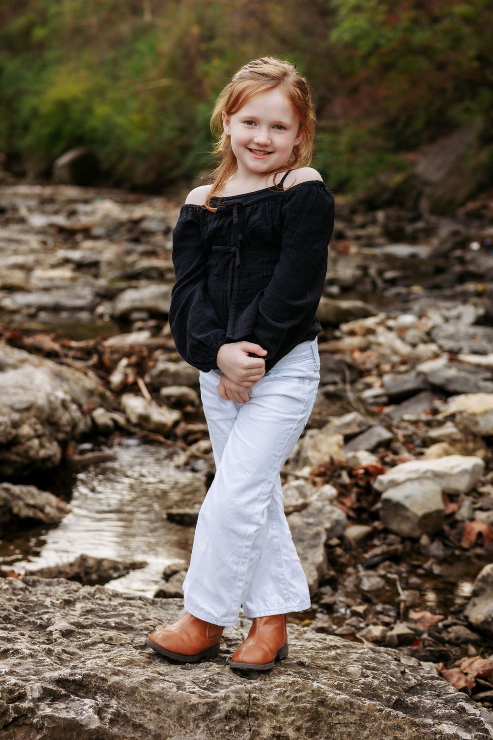 A young girl in a black top and white pants poses on rocks near a stream.
