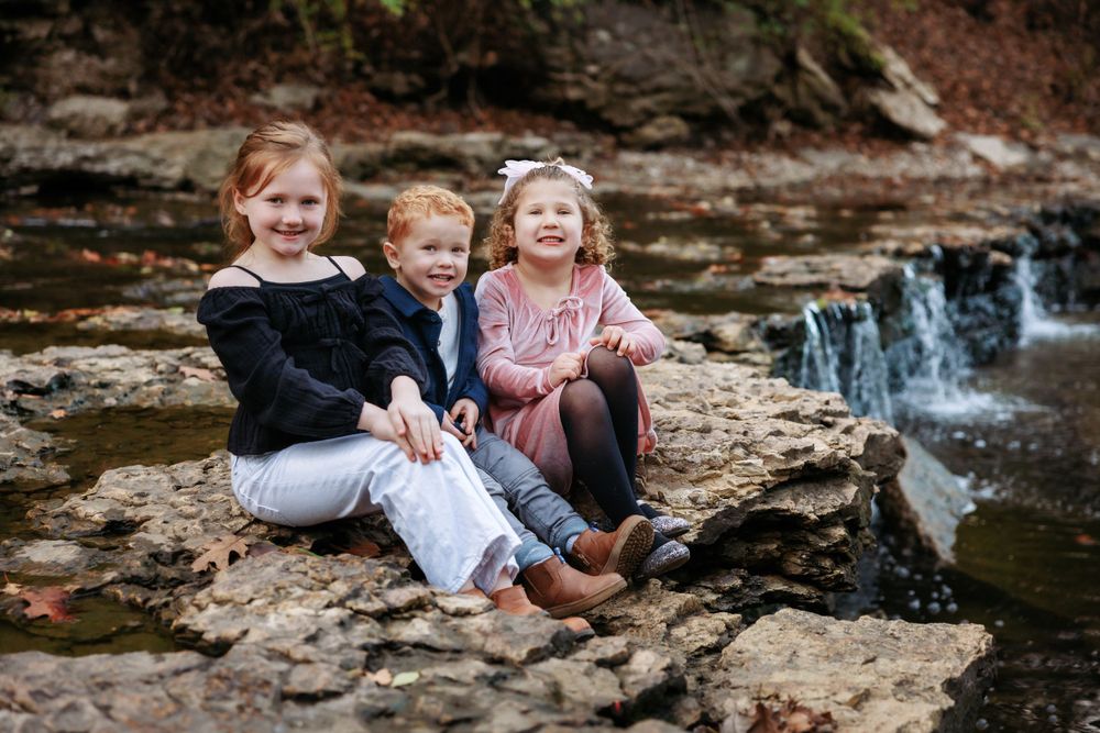 Three children sitting together on rocks by a small waterfall enjoying nature.