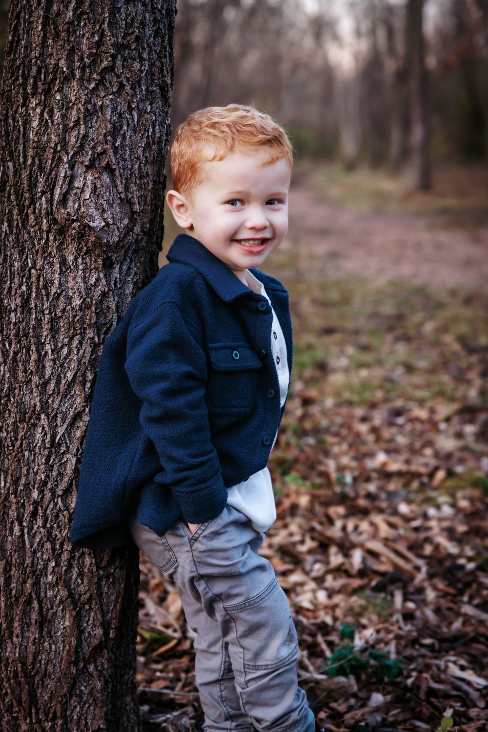 Child in navy jacket poses playfully against large tree trunk in autumn forest.