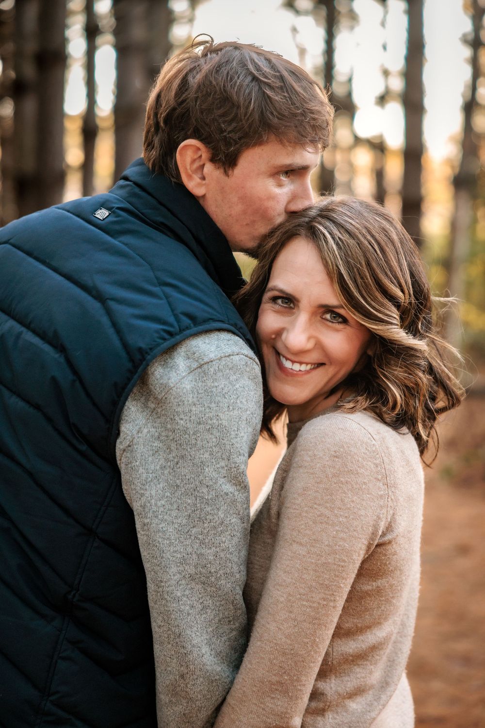 A tender moment between a couple in warm clothing against a backdrop of tall pine trees.