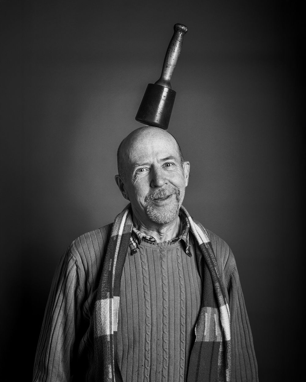 Black and white portrait of a person wearing a cable knit sweater and striped scarf with a bottle balanced on their head.