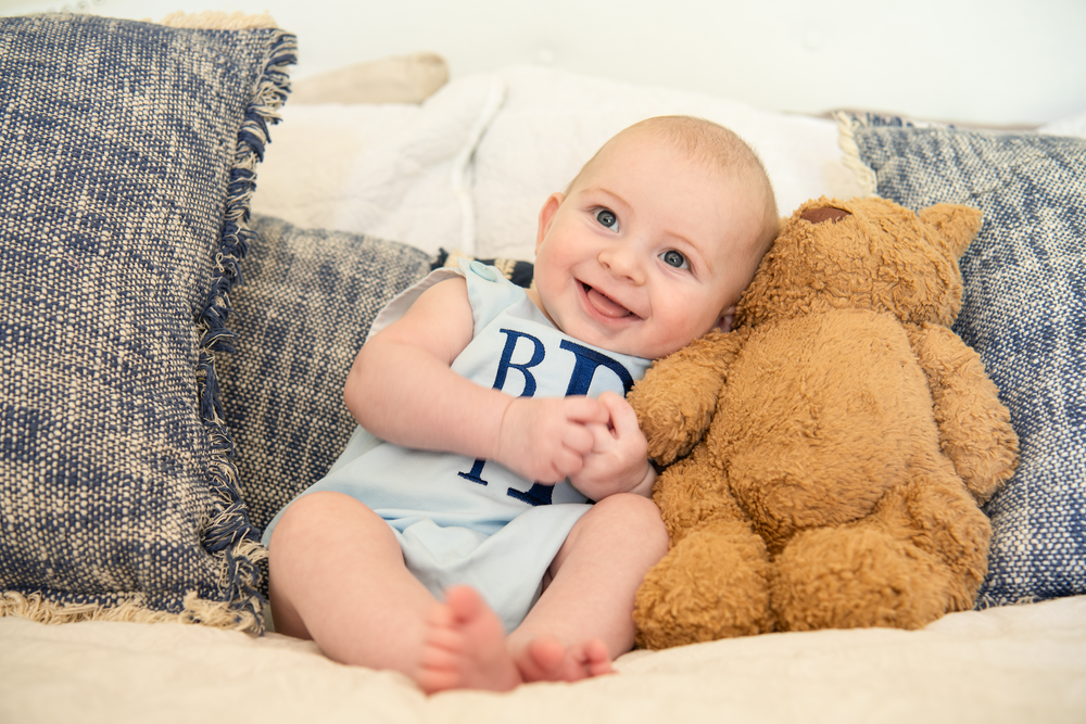 Cheerful baby boy in blue romper smiling beside a brown teddy bear on a white bed.