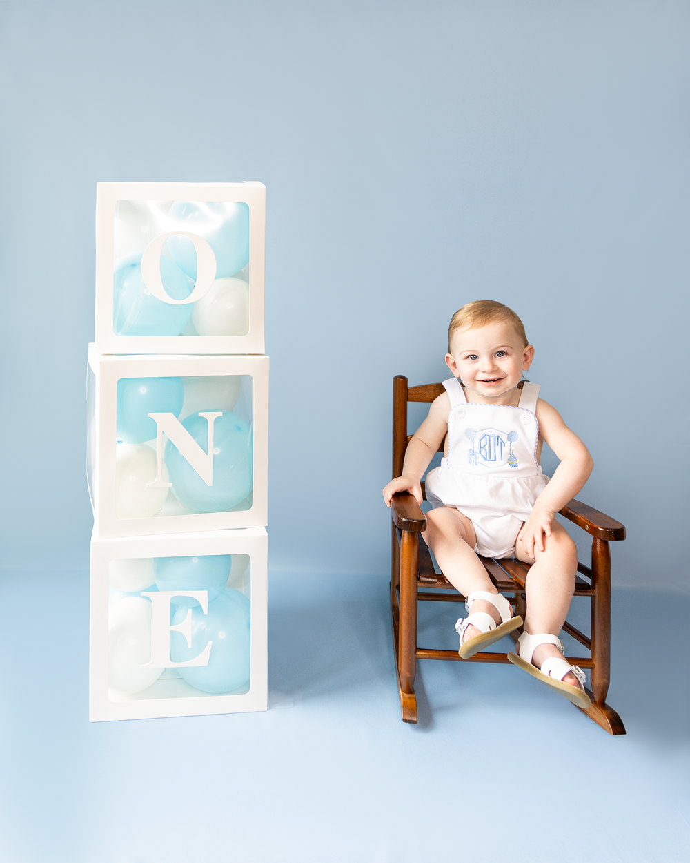 Baby boy in monogrammed romper sits smiling in wooden rocking chair beside stacked white ONE letter blocks.