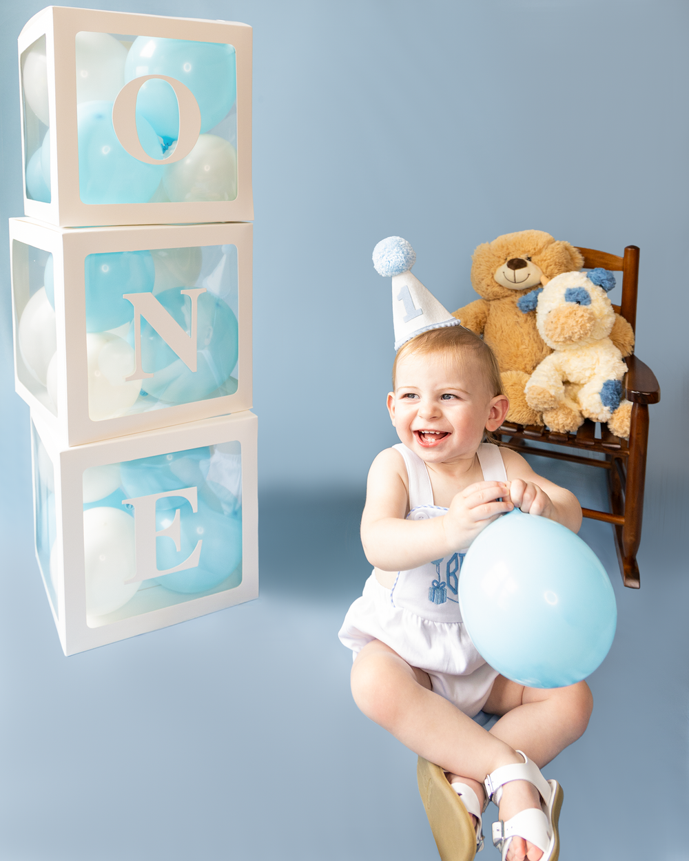 Laughing baby boy in party hat holds blue balloon seated in rocking chair with teddy bears beside ONE letter blocks.