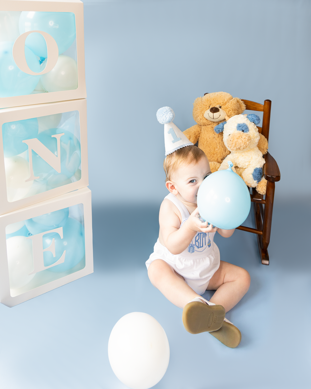 Baby boy in party hat sits with teddy bears and blue balloons beside ONE letter boxes for first birthday.
