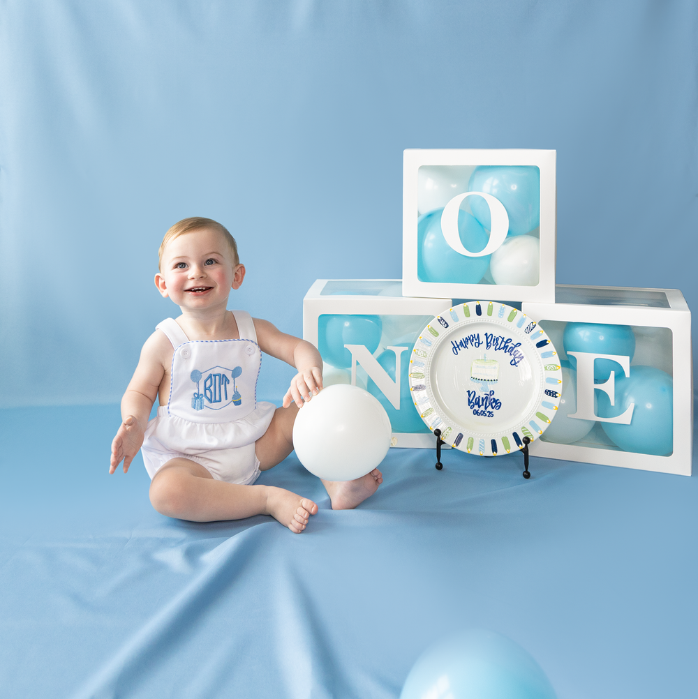Smiling baby boy poses with white balloon beside ONE letter boxes and birthday plate on blue backdrop.