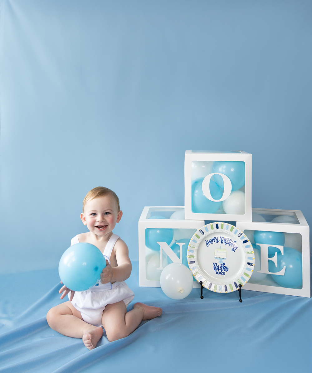 Smiling baby girl celebrates first birthday with blue balloons and ONE letter boxes on blue backdrop.