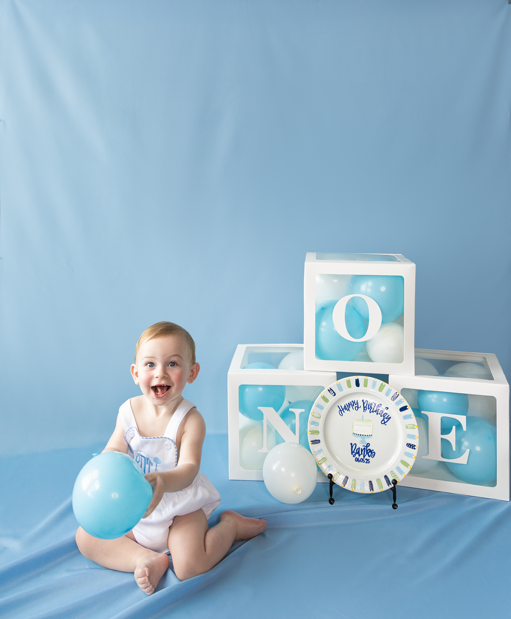 Laughing baby girl sits with blue and white balloons beside ONE letter boxes and birthday plate.