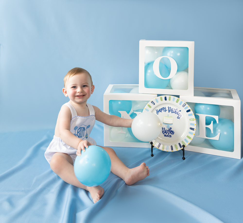 Happy baby boy smiles beside ONE letter boxes, birthday plate, and blue balloon on blue backdrop.