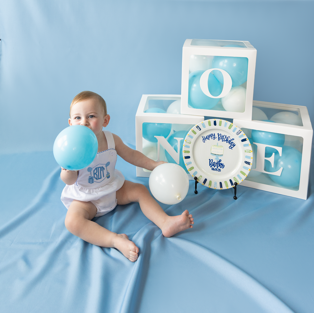 Baby boy blows up blue balloon beside ONE letter boxes and Happy Birthday plate at first birthday.