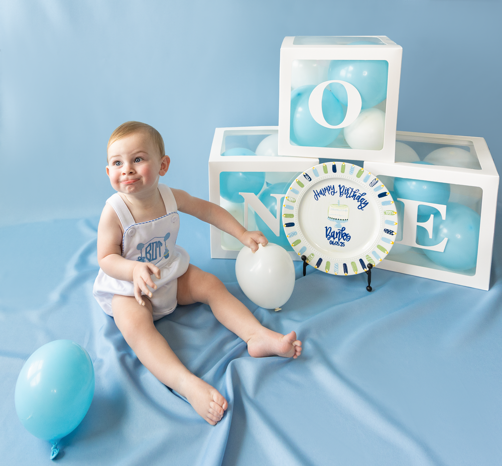 Cheerful baby boy sits with white balloon beside ONE letter boxes and birthday plate on blue backdrop.