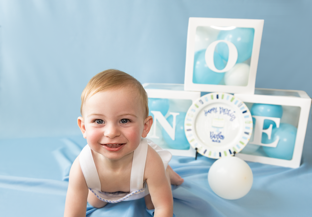 Close-up of smiling baby boy crawling in front of ONE letter boxes and birthday plate on blue backdrop.