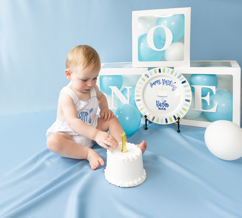 Baby boy smashes first birthday cake beside ONE letter boxes and Happy Birthday plate on blue backdrop.