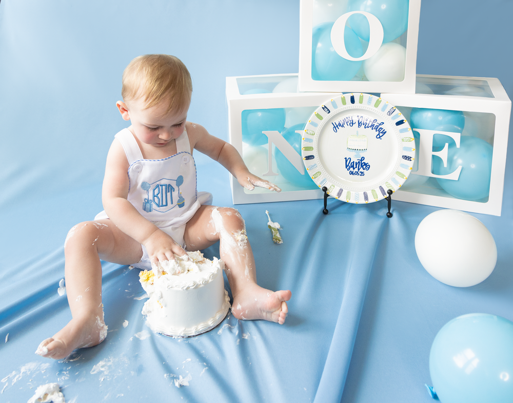 Baby boy smashes white birthday cake beside ONE letter boxes and Happy Birthday plate on blue backdrop.