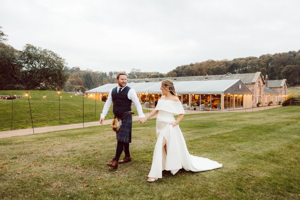 A couple in wedding attire walks through a scenic grassy field during their countryside celebration.