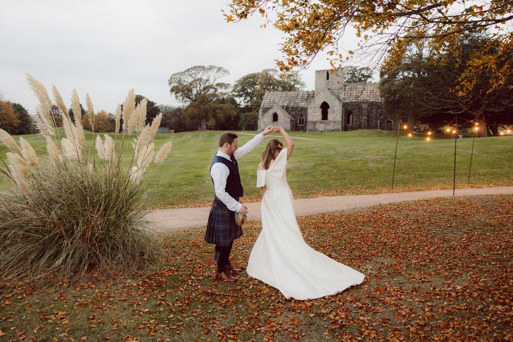 A couple in wedding attire walks through a scenic autumn landscape with fallen leaves and stone ruins in the background.