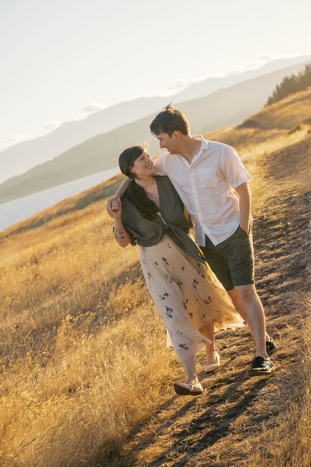 Two people share a tender moment on a sunlit hillside with mountains visible in the background during golden hour.