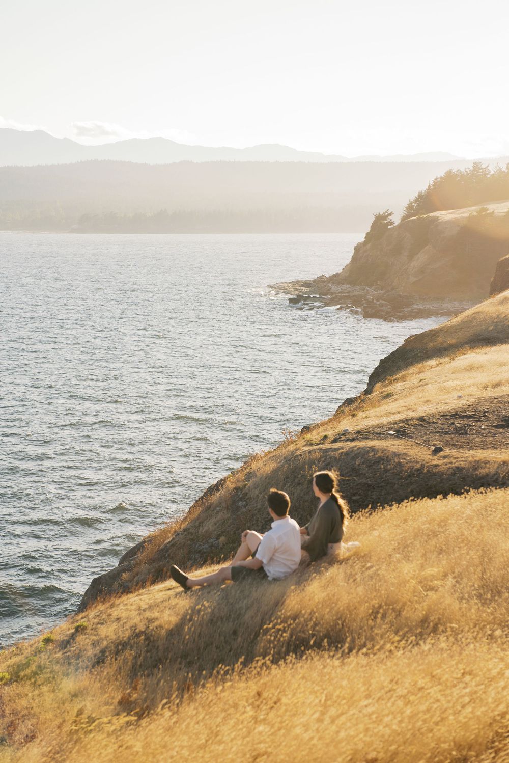 Two people sit together on a grassy coastal cliff overlooking the ocean at golden hour.