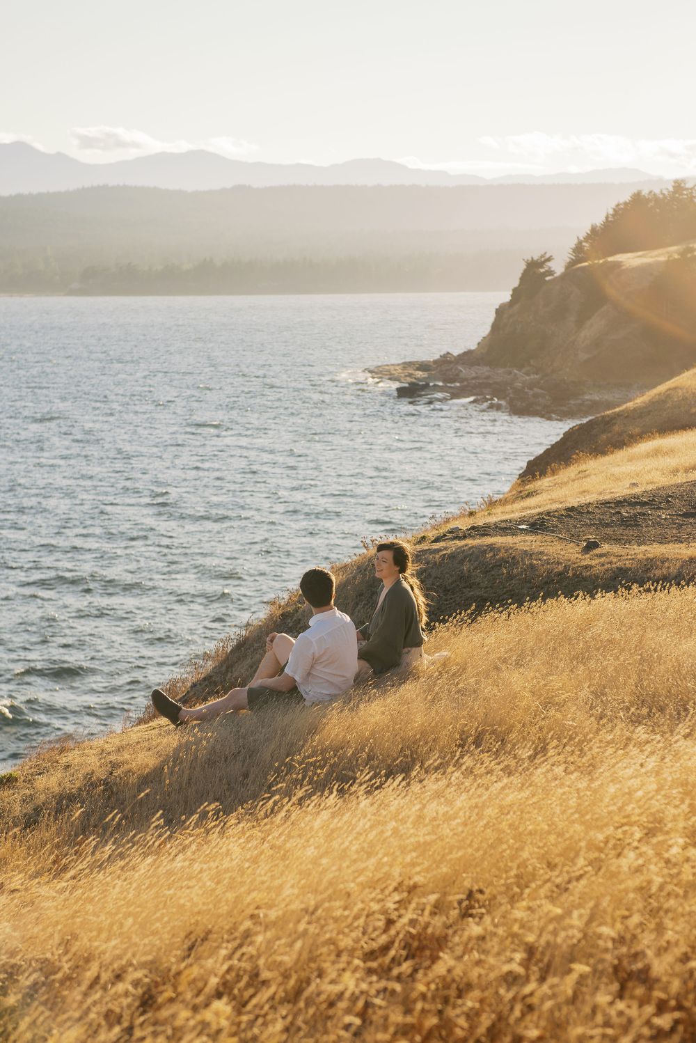 People sitting on grassy coastal cliff overlooking ocean and mountains during golden sunset on Pacific coast.
