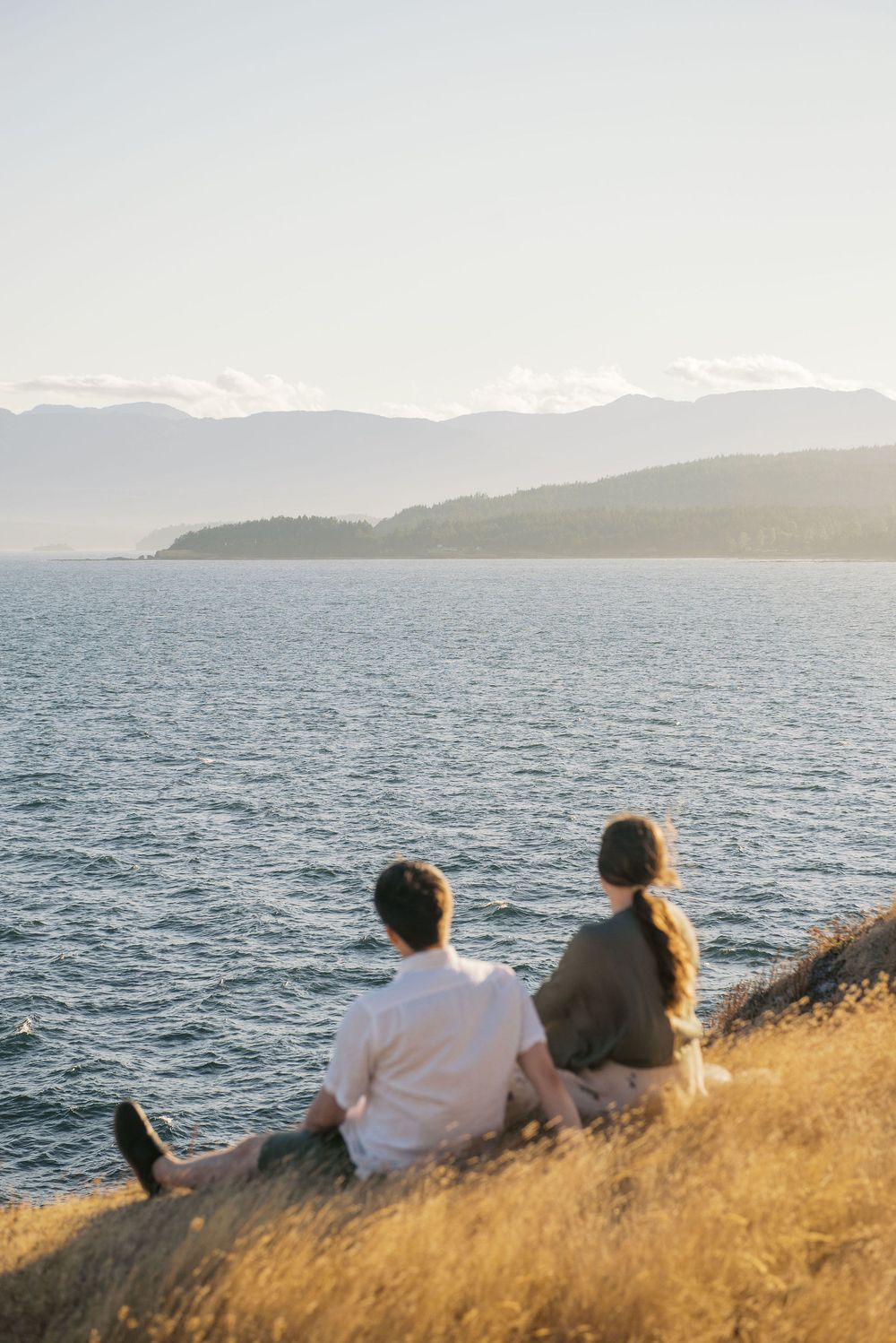 Two people sit on a grassy hillside overlooking a serene ocean view with mountains in the distance at sunset.