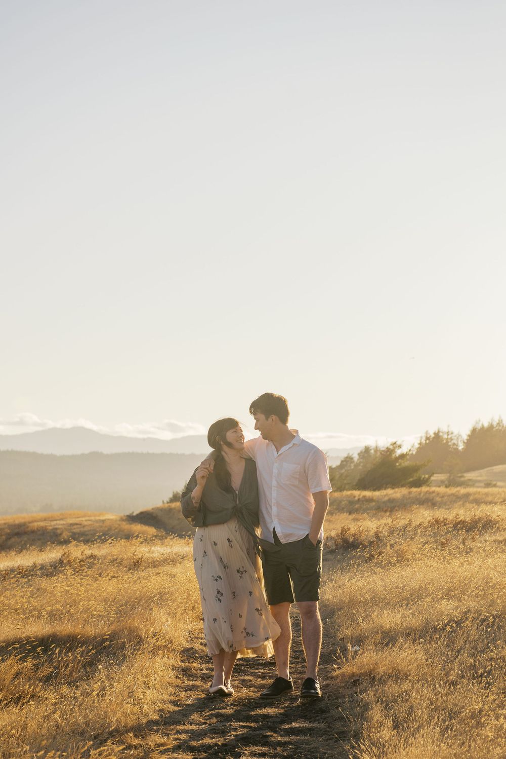 A couple shares a romantic moment in a golden sunlit meadow during sunset with mountains in the background.