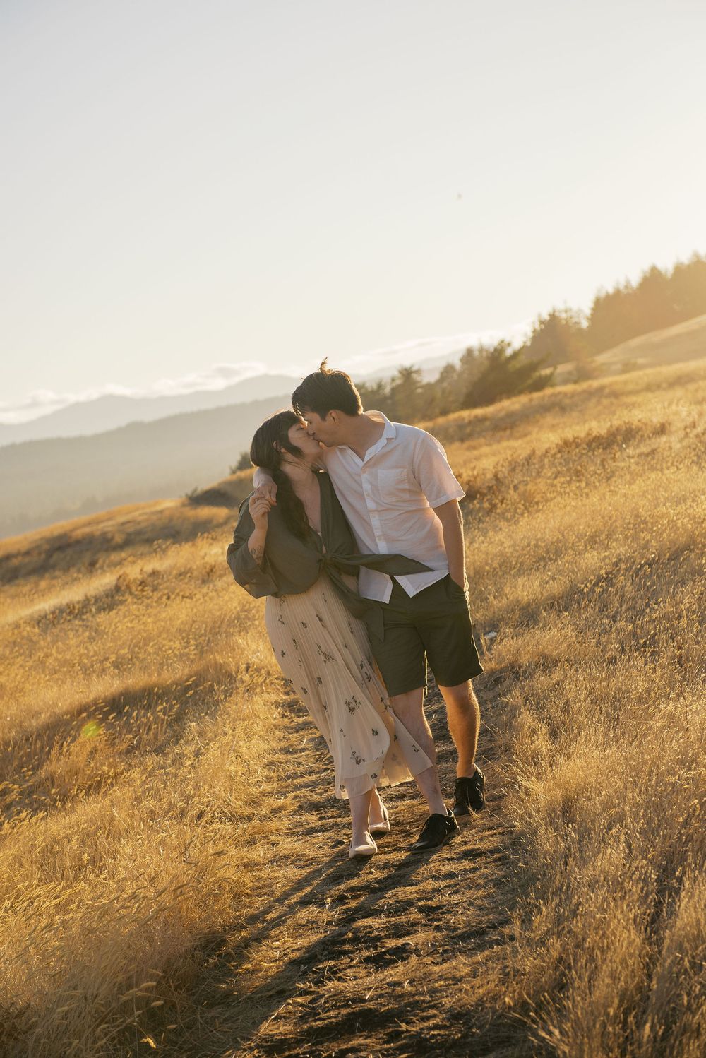 Couple embraces on a sunlit hillside during golden hour as warm light illuminates the dry grass slope.