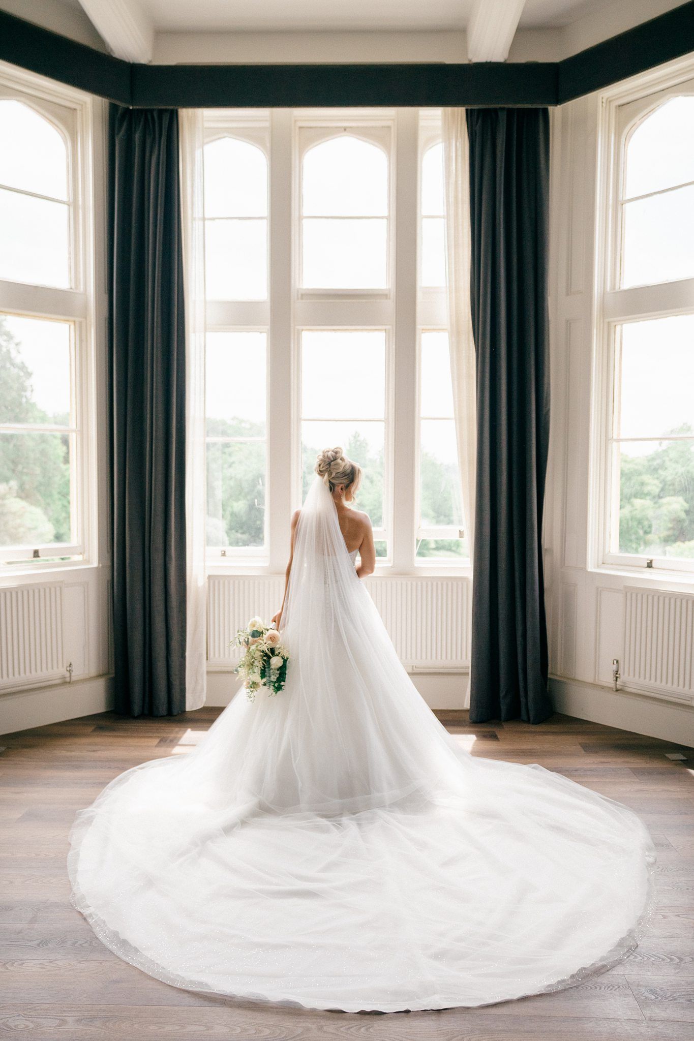 A flowing white wedding dress with a dramatic cathedral train displayed in a bright room with dark curtains and windows.