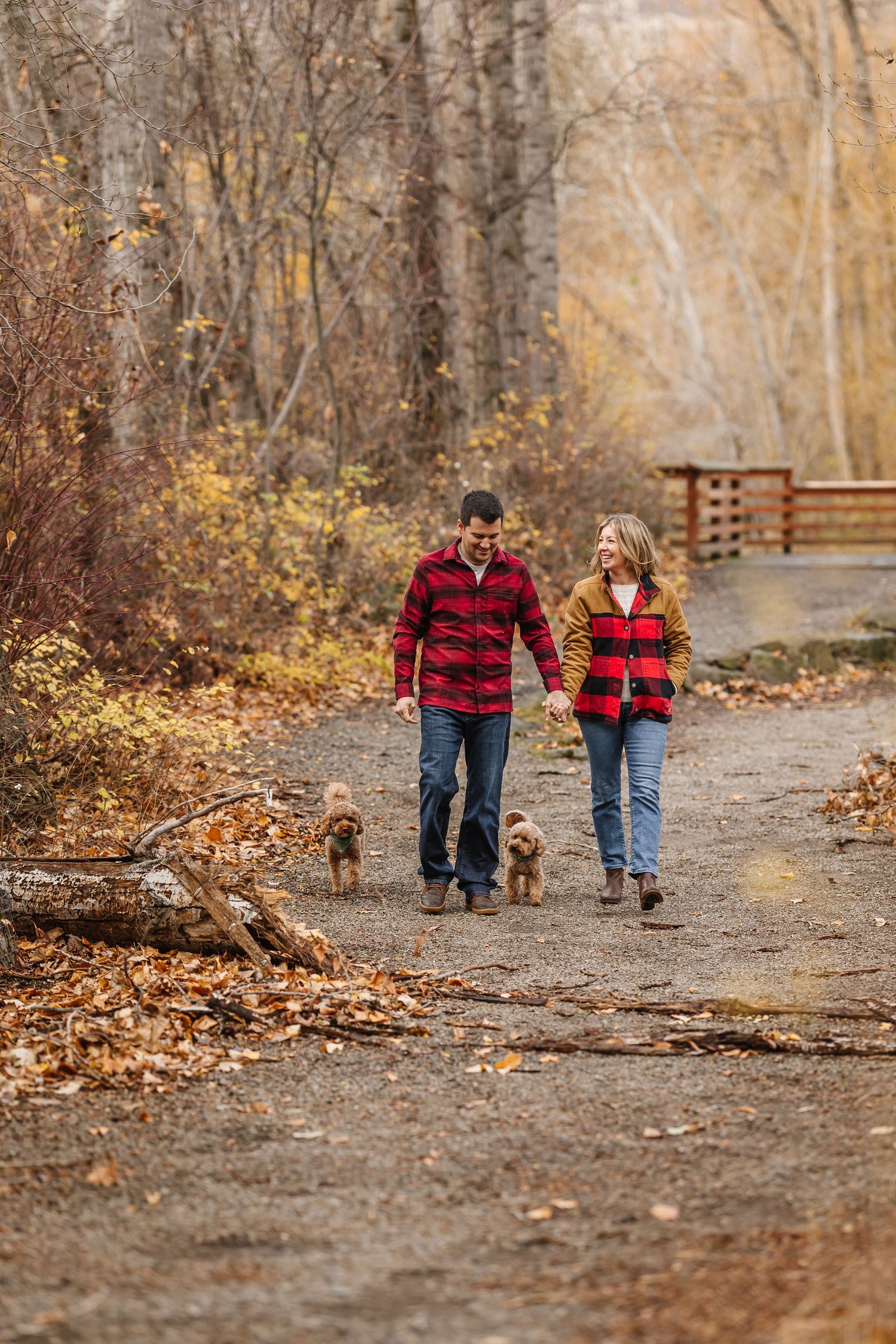 Two people in matching red plaid shirts walk hand in hand down a leaf-covered autumn trail with a bench in the distance.