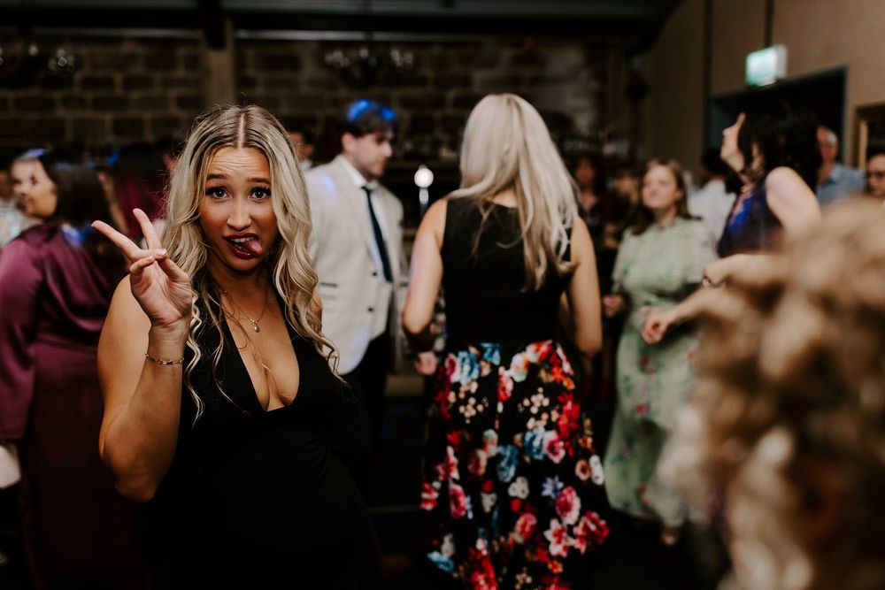 A woman in a black dress flashes a peace sign while enjoying a lively indoor party atmosphere.