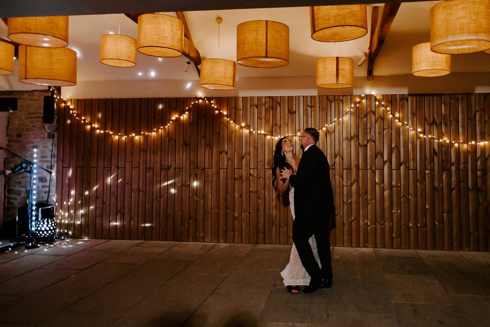 String lights illuminate a rustic barn-style venue interior with wooden walls and hanging drum shade pendant lights.