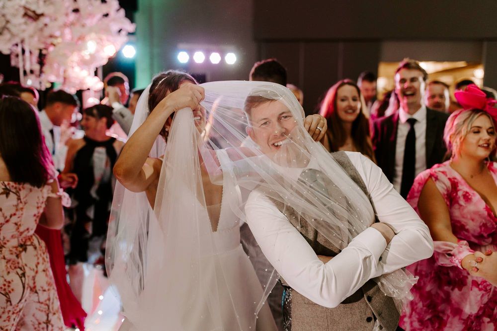 Wedding guests dance and celebrate on a lively dance floor illuminated by stage lights during a reception party.