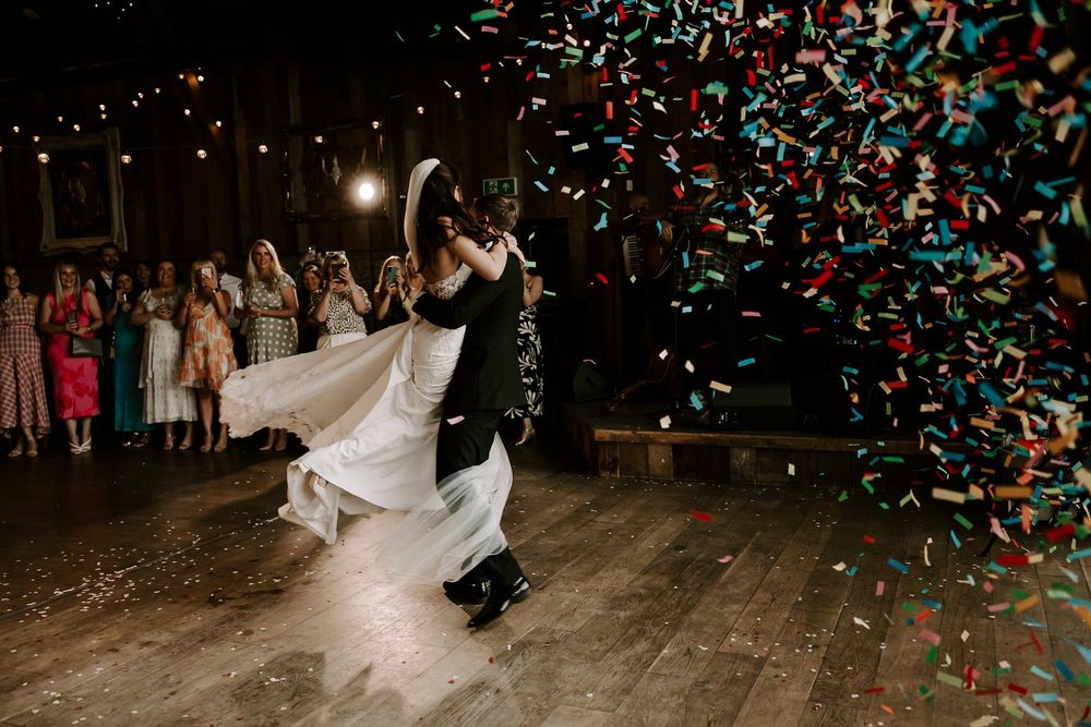 Wedding couple dances under falling confetti on a wooden dance floor during their first dance celebration.