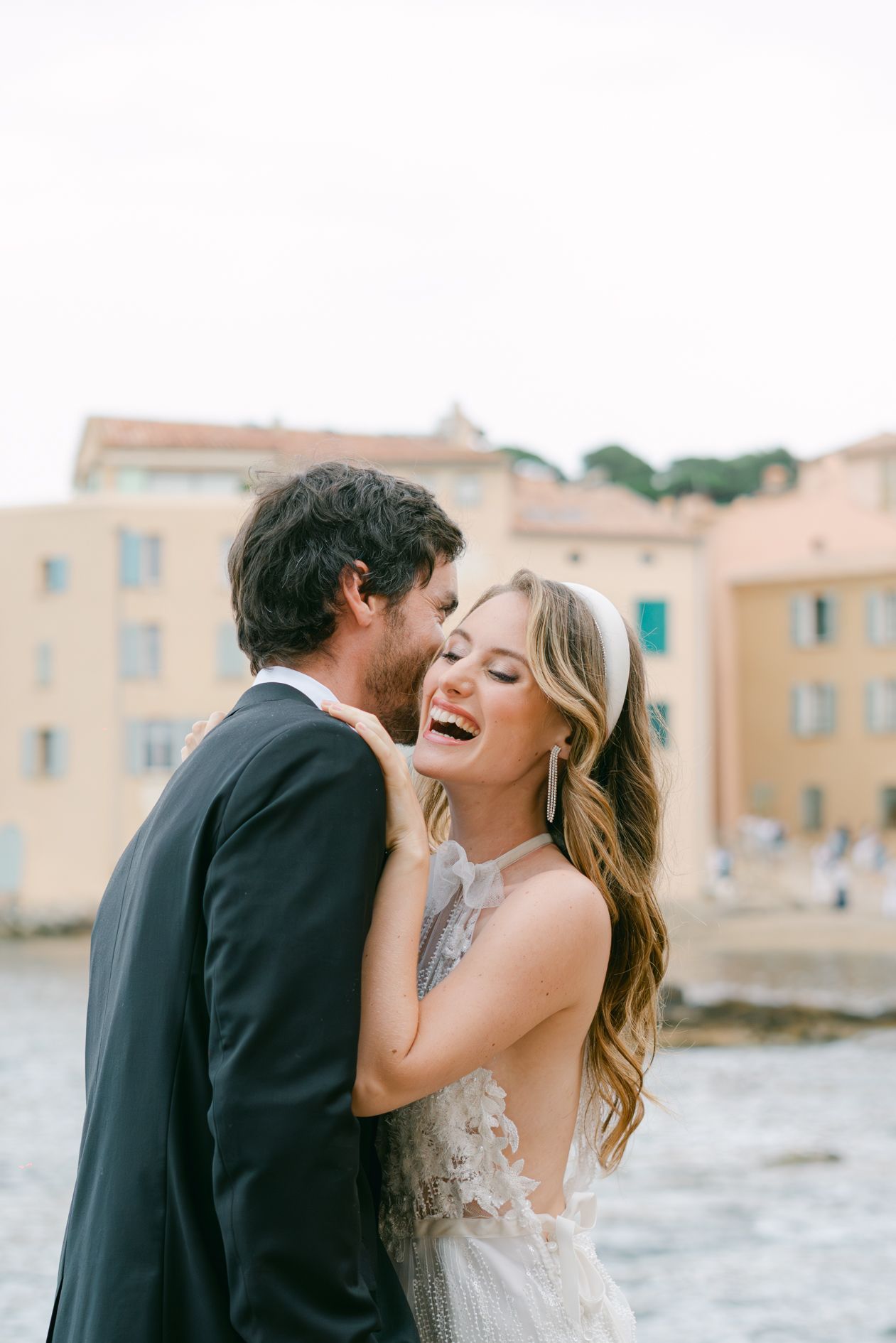 A joyful couple shares an intimate moment by the water in front of pastel Mediterranean buildings.