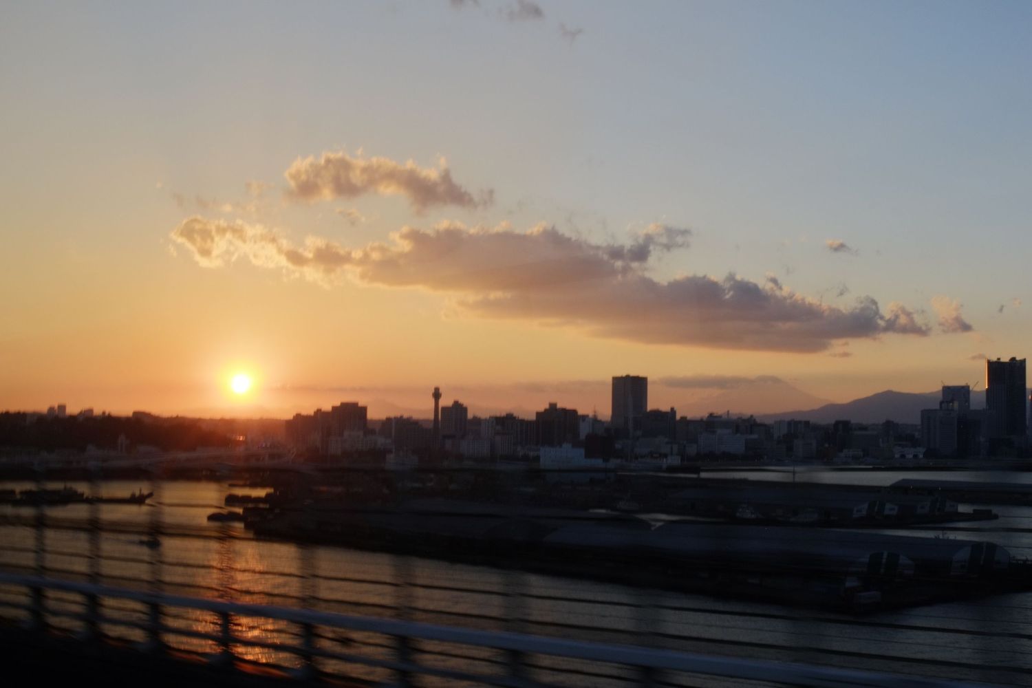Stunning sunset silhouettes city skyline and harbor with warm orange glow reflecting off clouds and water.