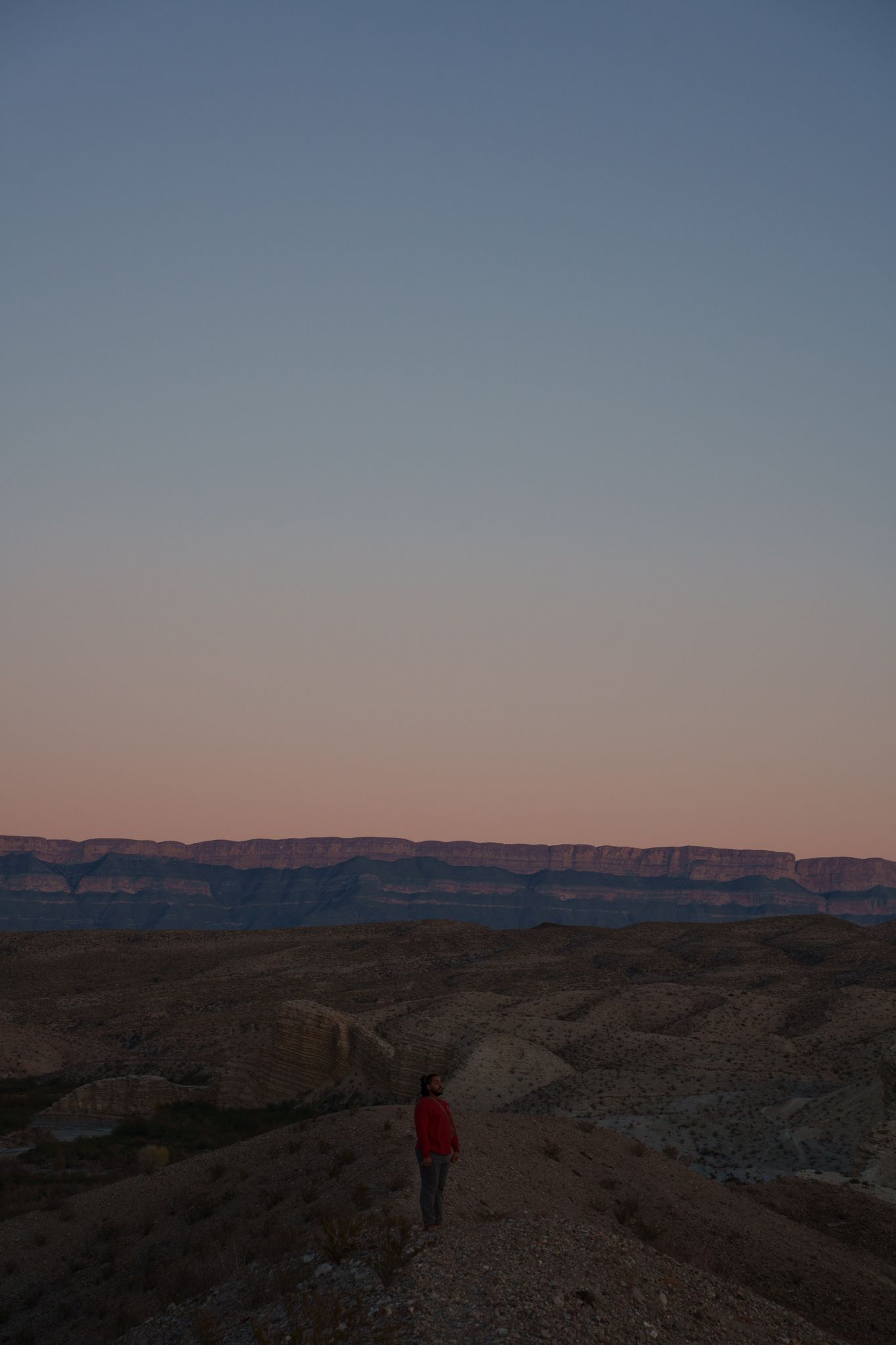 A person in a red jacket stands on a desert trail at sunset with distant cliffs in the background.