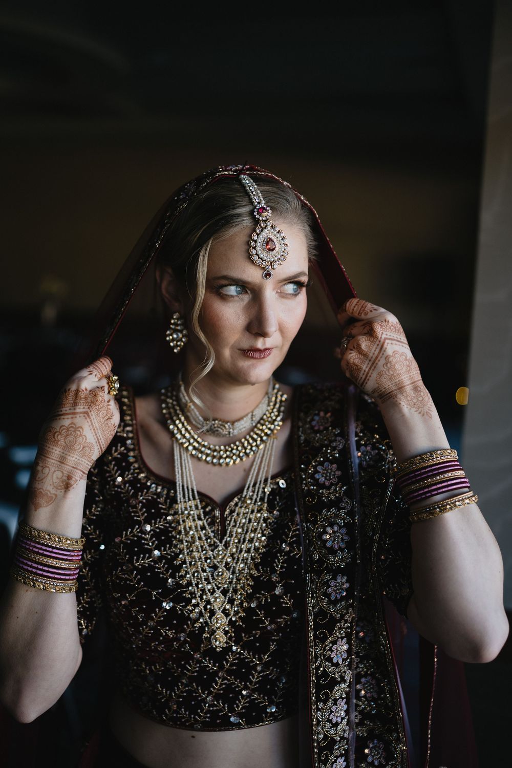 Bride in black embroidered lehenga with gold jewelry and maang tikka adjusts veil in dim lighting.at Sandia Resort and Casino