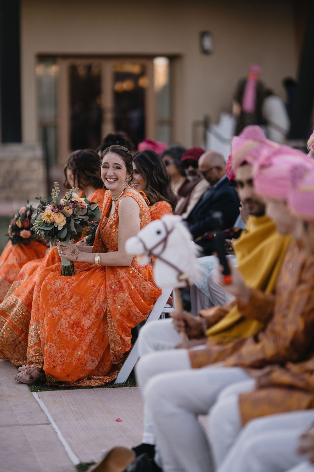 Smiling bridesmaid in orange lehenga holds bouquet while seated at outdoor Indian wedding ceremony.at Sandia Resort and Casino