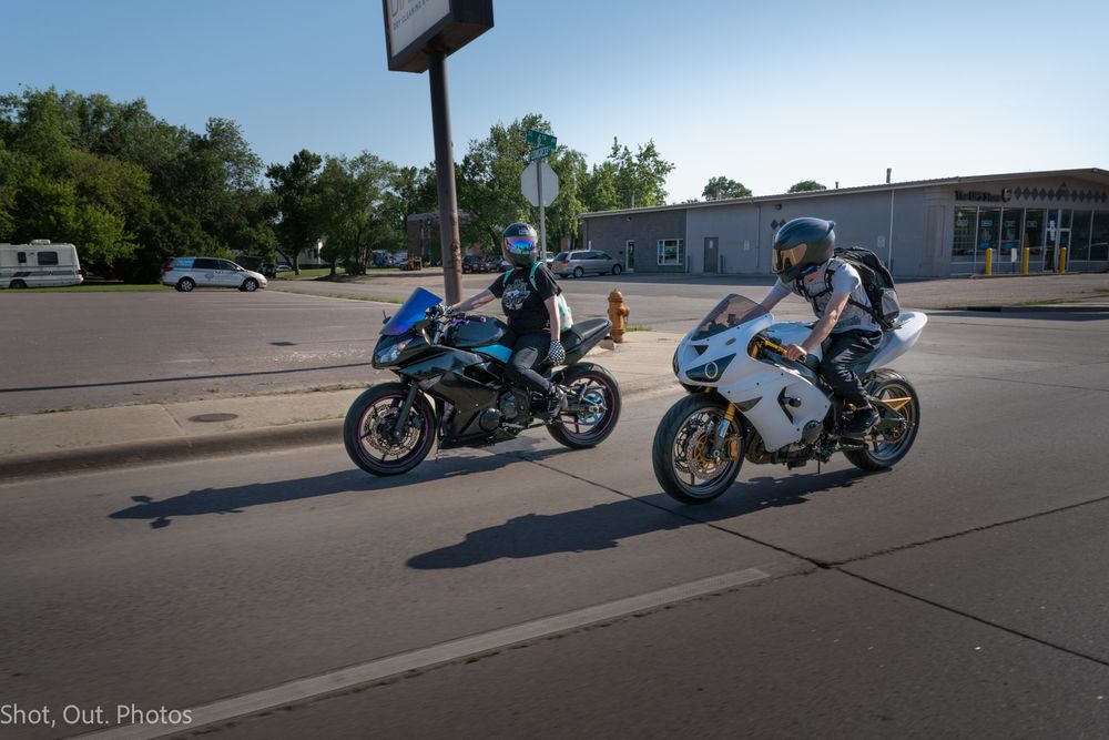 Two sport motorcycles ridden by helmeted riders on a sunny urban street with trees and buildings in the background.