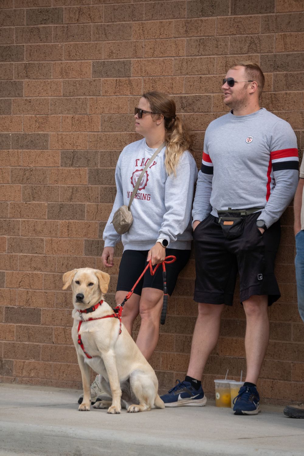 Couple with yellow Labrador on red leash standing against brick wall, wearing casual sweatshirts and shorts outdoors.