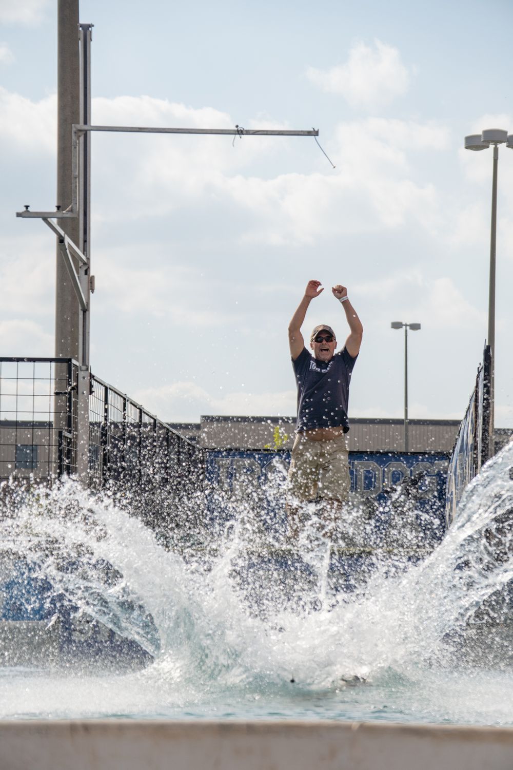 Man with arms raised triumphantly behind splashing water fountain outdoors on a cloudy day.