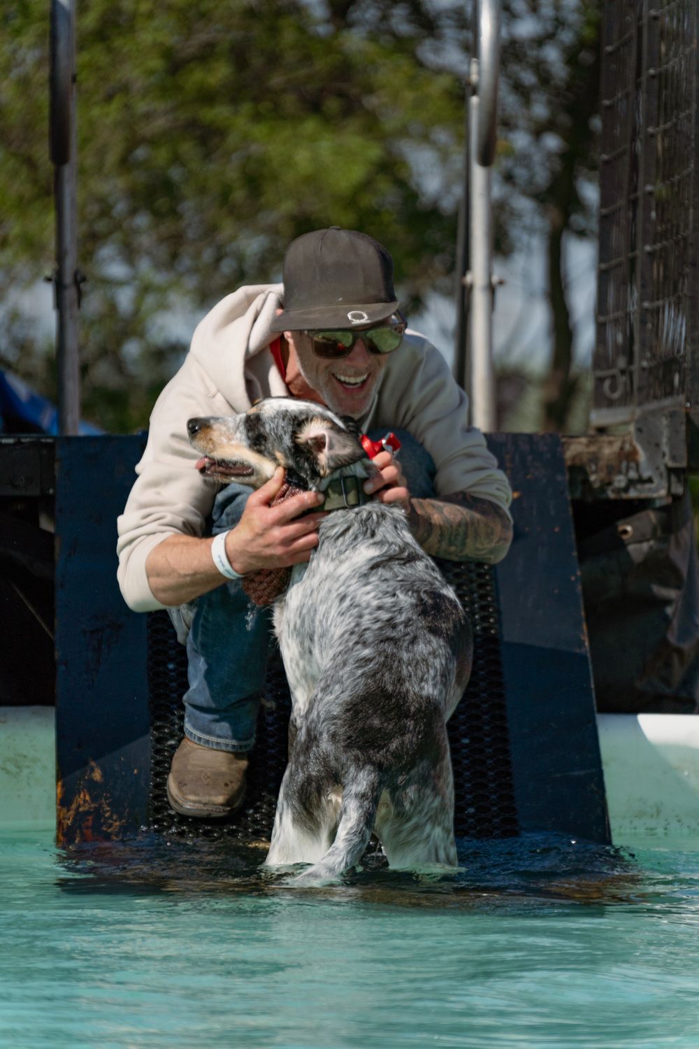 Man in cap and sunglasses rewarding a wet blue heeler dog standing in a shallow pool outdoors.