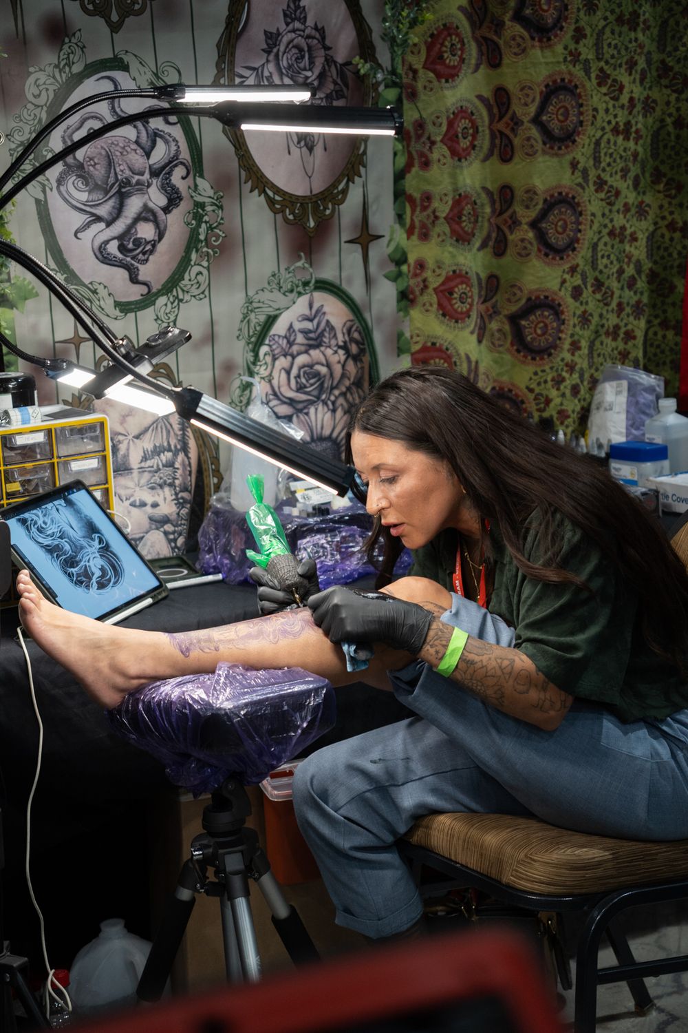 Female tattoo artist in black gloves tattooing a client's leg at a tattoo convention booth with artwork on walls.